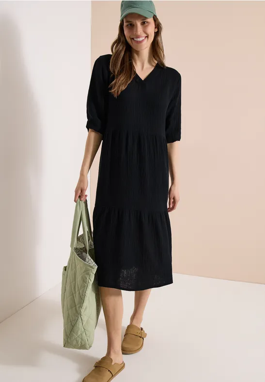 Femme en robe midi noire à encolure en V et jupe à volants, porte un sac en tissu vert et des chaussons.