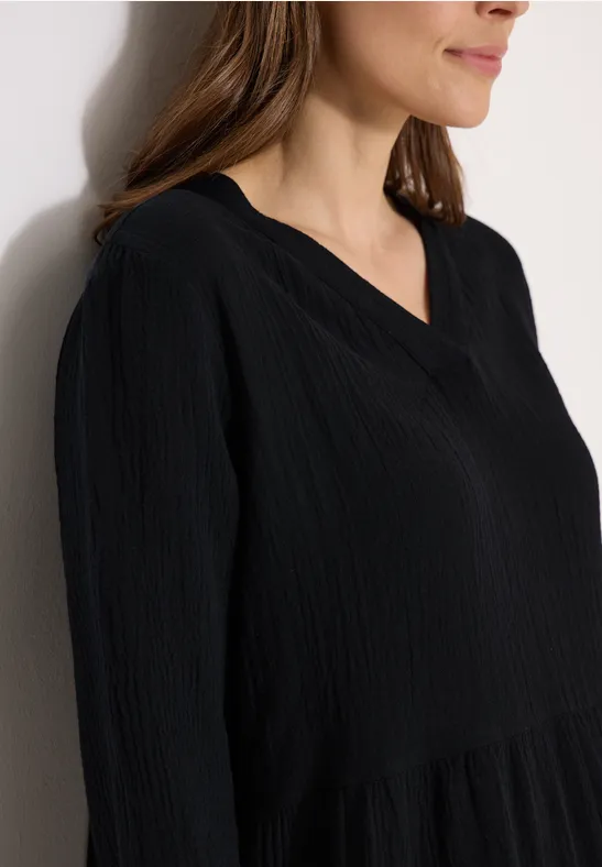 Femme en gros plan portant un chemisier noir à texture légère et col en V, appuyée contre un mur clair.