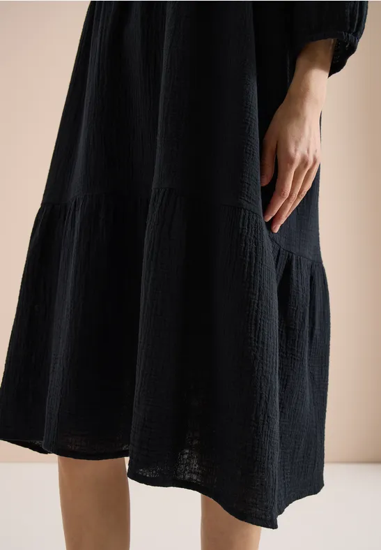 Gros plan sur une robe midi noire texturée, coupe ample, main pendante visible devant un fond clair.