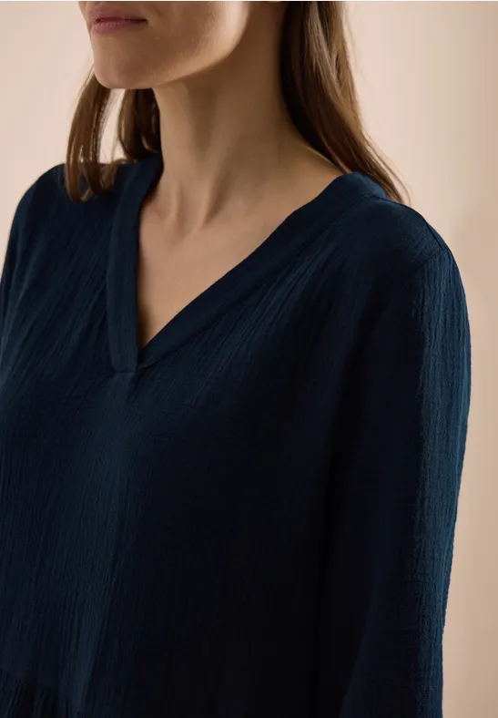Eine Frau trägt eine dunkelblaue, strukturierte V-Ausschnitt-Bluse vor hellem Hintergrund.