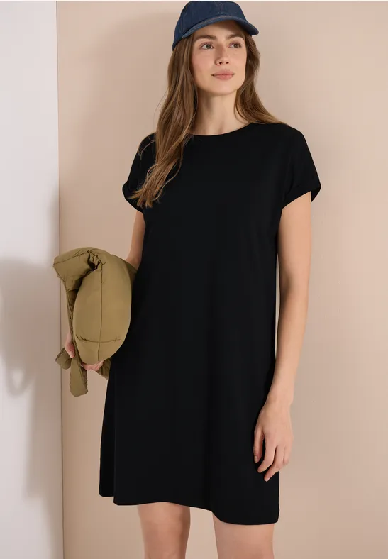 Frau in schwarzem T-Shirt-Kleid mit Kapuze hält eine grüne Tasche vor beiger Wand.
