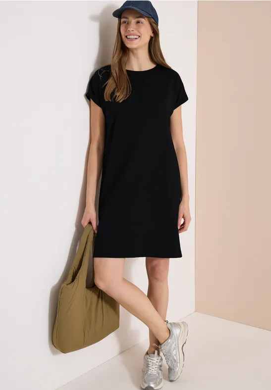 Frau in schwarzem T-Shirt-Kleid, Baseballcap, weißen Sneakers und großer olivgrüner Tasche.