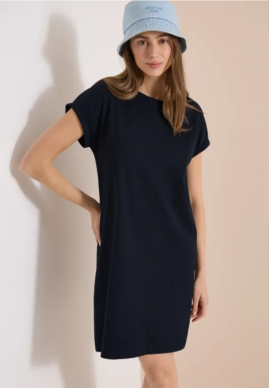 Eine Frau in lässigem marineblauen T-Shirt-Kleid und hellem Anglerhut vor neutralem Hintergrund.