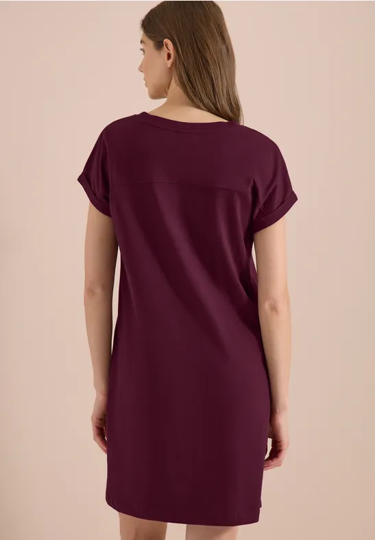 Femme de dos portant une robe bordeaux ample mi-longue à manches courtes sur fond beige