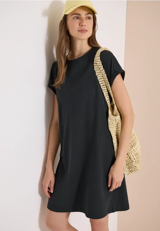 Eine Frau trägt ein schwarzes T-Shirt-Kleid und hält eine geflochtene Strohtasche vor hellem Hintergrund.