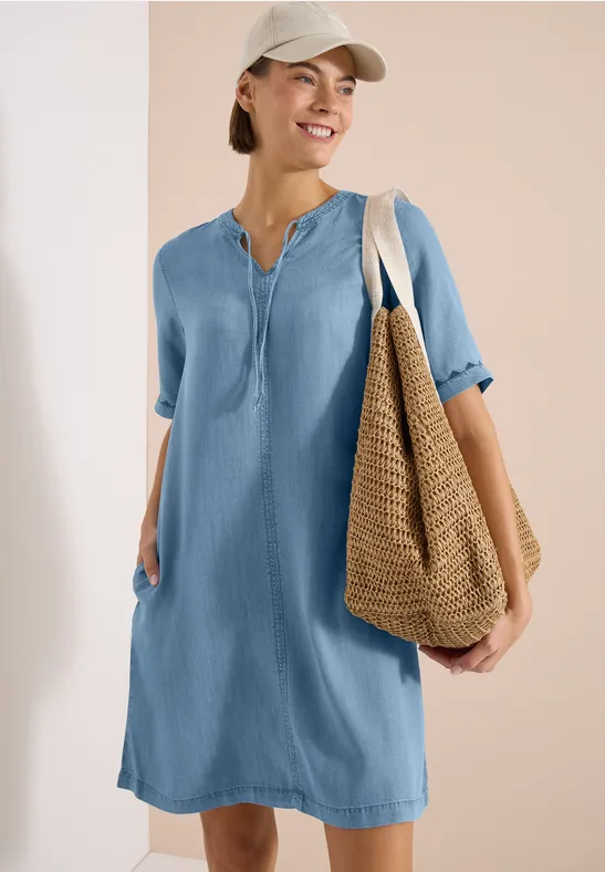 Femme en robe-chemise bleu clair décontractée, manches retroussées, grand sac en paille et casquette