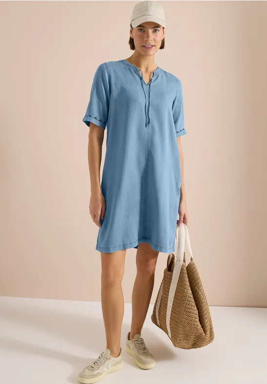 Femme en robe bleu clair décontractée, baskets blanches et sac en paille, posant devant un fond neutre.