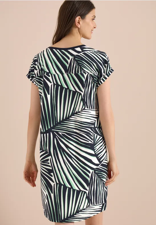 Femme de dos portant une robe à manches courtes à imprimé feuilles de palmier bleu-vert sur fond beige.