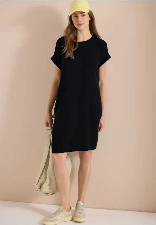 Frau in schwarzer T-Shirt-Kleid, Sneakers und Tasche vor neutralem Hintergrund.