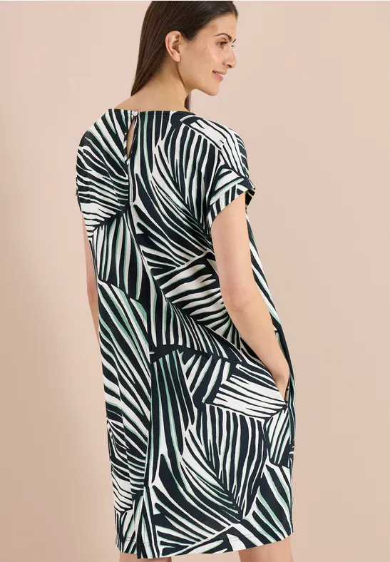 Femme sur fond beige, vêtue d'une robe ample à manches courtes à motif de feuilles vertes et blanches avec poches.