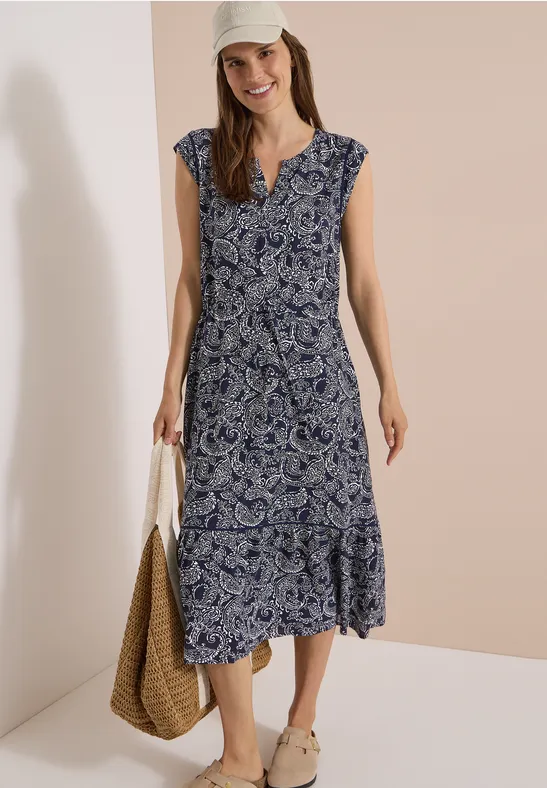 Mannequin porte une robe midi sans manches à motif paisley bleu et blanc, décolleté en V, sac en osier et sandales.
