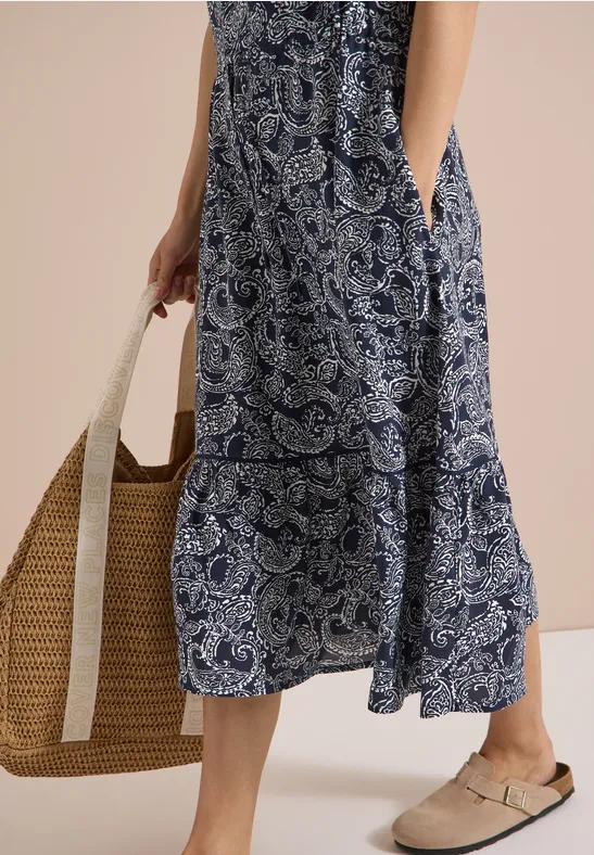Femme portant une robe longue à motif cachemire bleu-blanc, un sac en paille et des mules confortables.