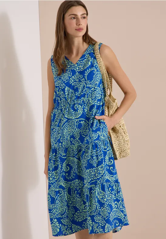 Femme en robe sans manches à motif paisley bleu-vert, col en V, avec sac tressé, fond uni.