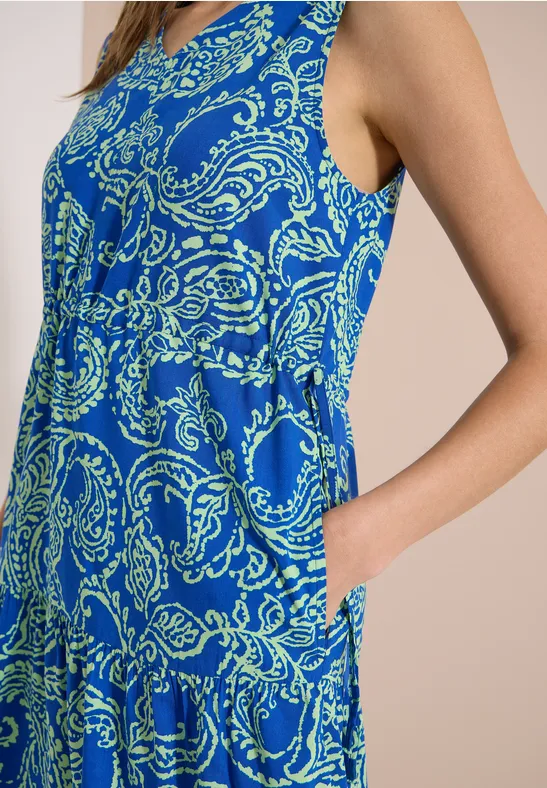 Femme de profil en robe sans manches bleue à motif cachemire, main dans la poche.