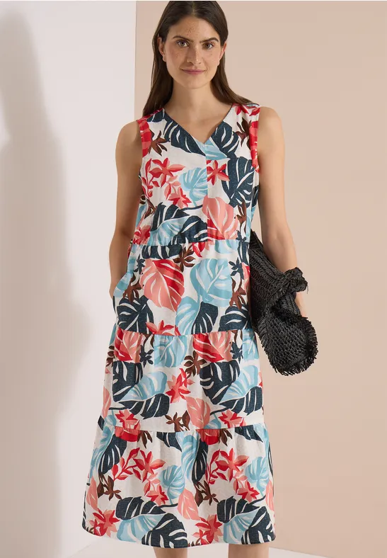 Frau in ärmellosem, gestuftem Maxikleid mit bunter Blumenoptik, dazu eine schwarze Netztasche.
