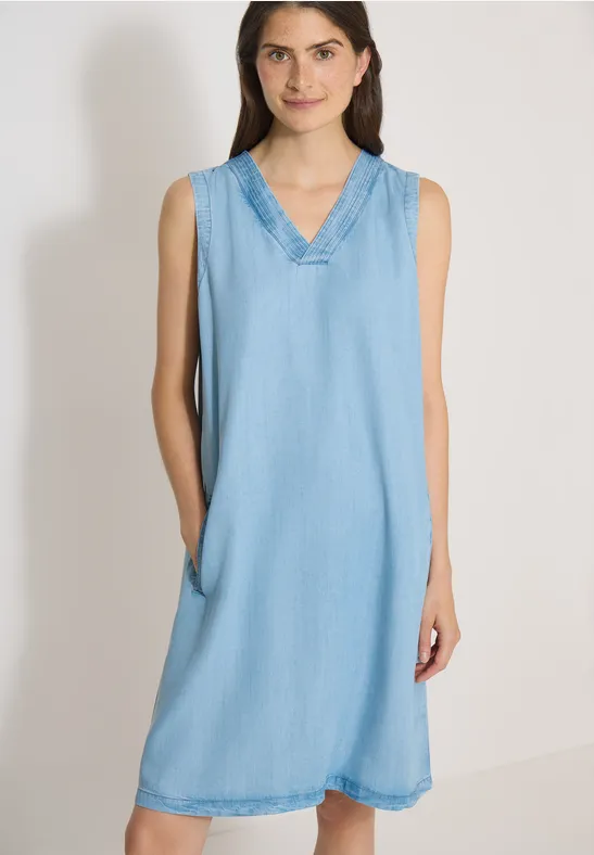 Femme porte une robe bleue claire sans manches, col en V, longueur genou et poches latérales.