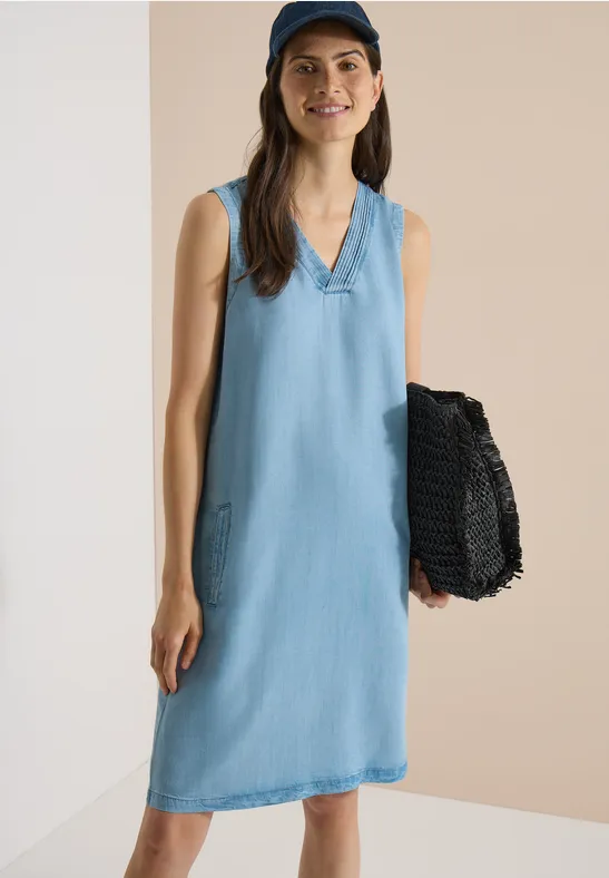 Femme en robe en jean bleu clair sans manches, décolleté en V, tenant un panier noir tressé devant un mur beige.