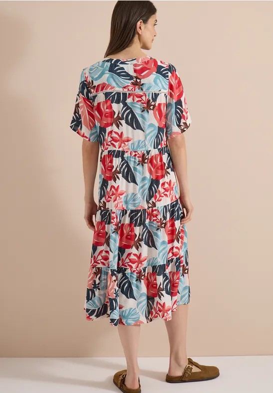 Rückansicht einer Frau in weißem, knielangem Kleid mit buntem Blumenmuster und weiten Ärmeln vor neutralem Hintergrund.