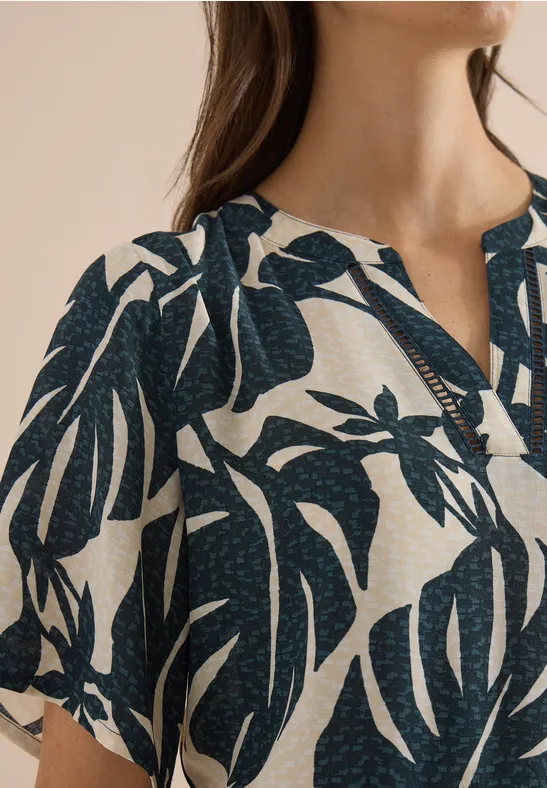 Nahaufnahme eines Oberkörpers in weißer Bluse mit dunkelblauem Monstera-Blattmuster.