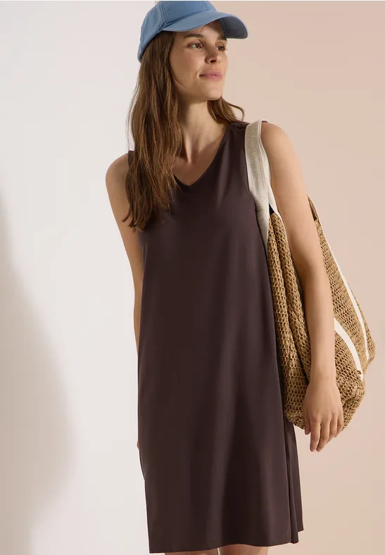 Frau in lässigem, ärmellosem Sommerkleid mit Strohtasche und Baseballcap vor neutralem Hintergrund.