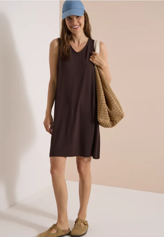 Model trägt ärmelloses, locker geschnittenes, dunkles Kleid, geflochtene Tasche in der Hand, helle Slip-on-Schuhe.