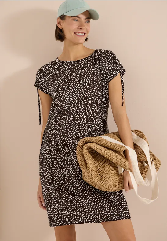 Frau in gemustertem Kurzarmkleid, mit strohfarbener Strandtasche und Baseballcap.