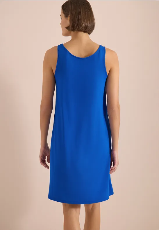Rückansicht einer Frau in einfachem, ärmellosem blauem Kleid vor einfarbigem Hintergrund.