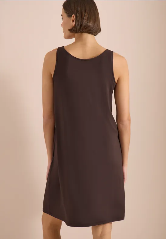 Eine Frau von hinten in einem ärmellosen braunen Kleid vor hellem Hintergrund.