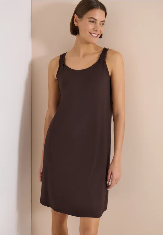 Eine Frau in einem schlichten, braunen, ärmellosen Trägerkleid steht vor hellem Hintergrund.