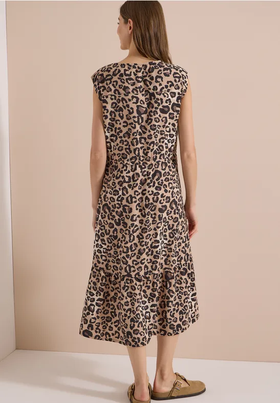 Frau von hinten in ärmellosem, midilangen Leopardenkleid und flachen Ballerinas vor beigem Hintergrund.