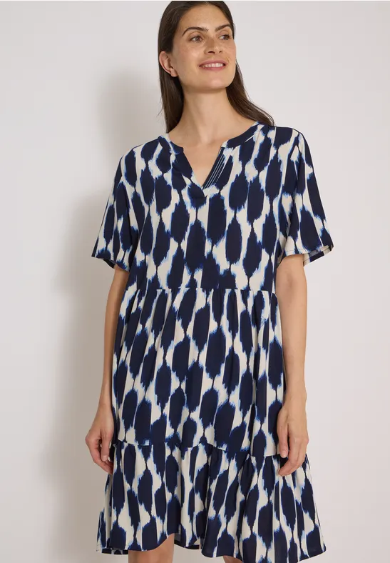 Femme porte une robe midi à manches courtes motif bleu et blanc, encolure en V et jupe à volants.