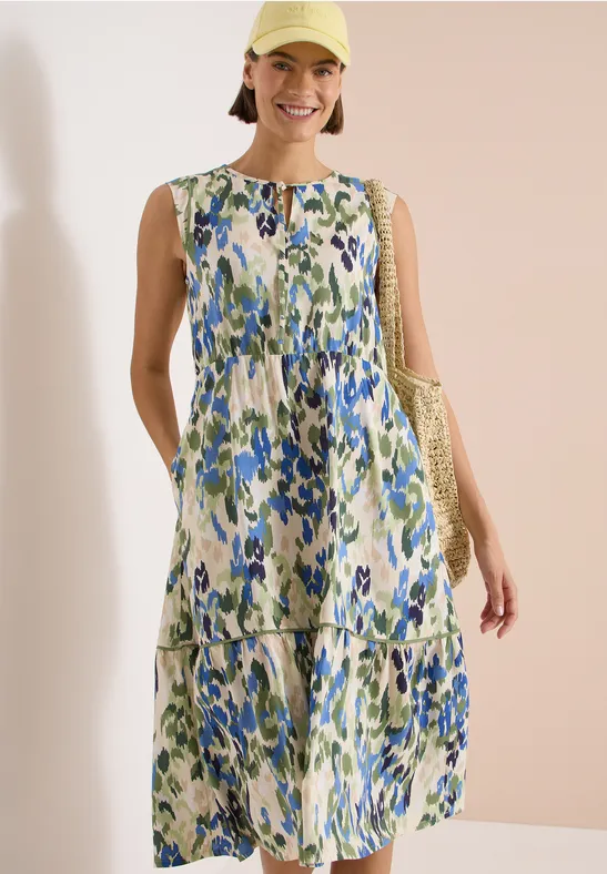 Frau in ärmellosem, blau-grünen, geblümtem Maxikleid, hält eine Tasche vor beigem Hintergrund.