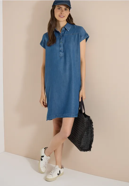 Femme en robe chemise en jean, baskets blanches, sac noir sur fond beige.