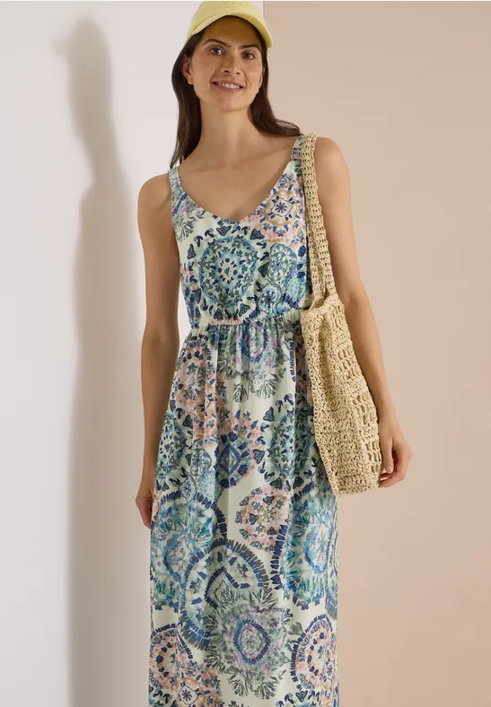 Eine lächelnde Frau trägt ein bunt gemustertes, ärmelloses Maxikleid, eine Strohtasche und eine gelbe Mütze.
