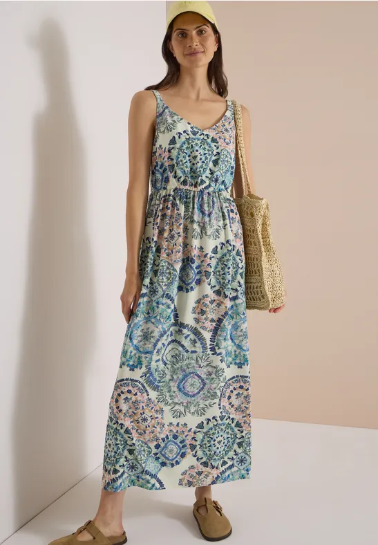 Frau im ärmellosen Maxikleid mit blau-grünem Fliesenmuster, Strohtasche und flachen Sandalen.