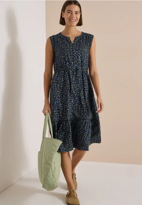 Frau in ärmellosem, knielangem dunkelblauem Blumenkleid mit Gürtel, trägt eine grüne gesteppte Einkaufstasche.