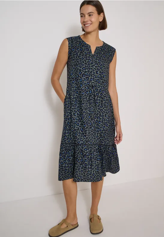 Frau in ärmellosem, dunkelblauem Midikleid mit kleinem Muster, gestufter Rockpartie und beigen Loafern.
