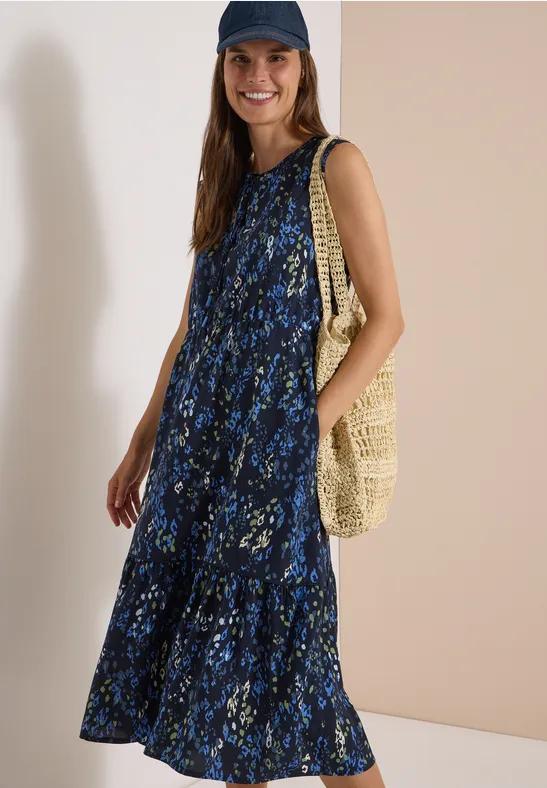 Frau in blauem geblümtem Maxikleid, mit Schirmmütze und Basttasche lächelt
