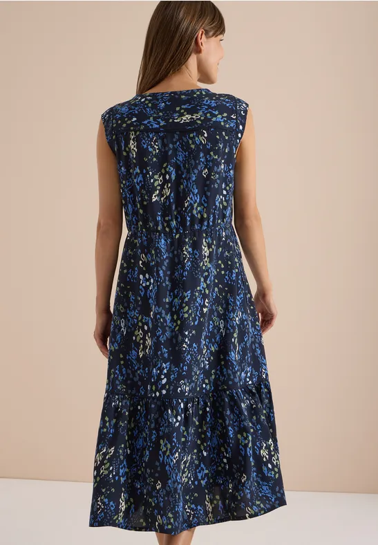 Eine Frau von hinten in einem blauen, ärmellosen, midilangen Kleid mit abstraktem Blumenmuster.
