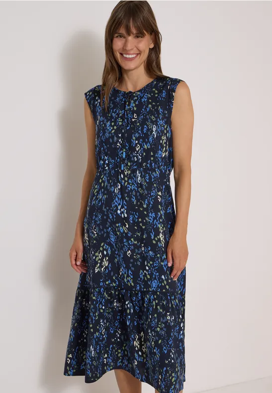 Frau in ärmellosem, bodenlangem Kleid mit blauem Blumenmuster vor hellem Hintergrund.