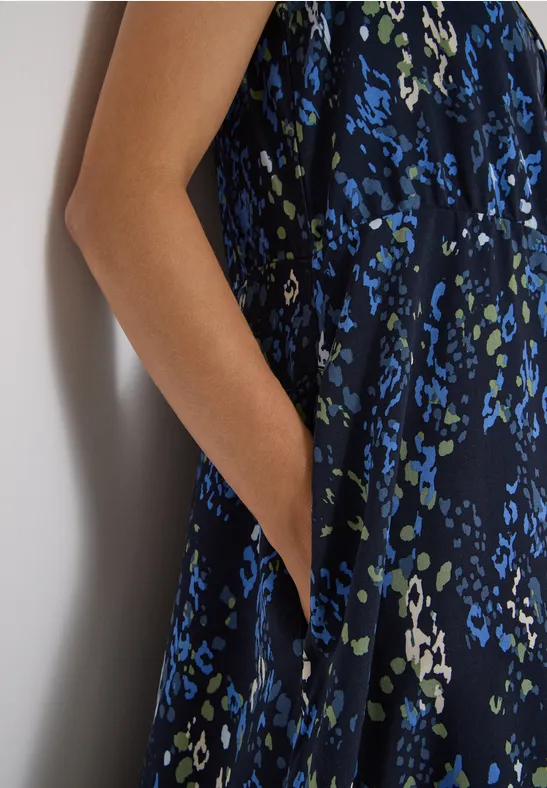 Ärmelloses, dunkelblaues Kleid mit blau-grünem Blumenmuster, Person stützt Arm an Wand, Hand in Tasche.