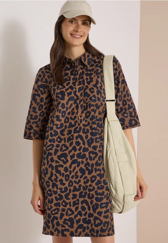 Femme souriante en robe léopard marron-noir à manches mi-longues, casquette et grand sac cabas crème.