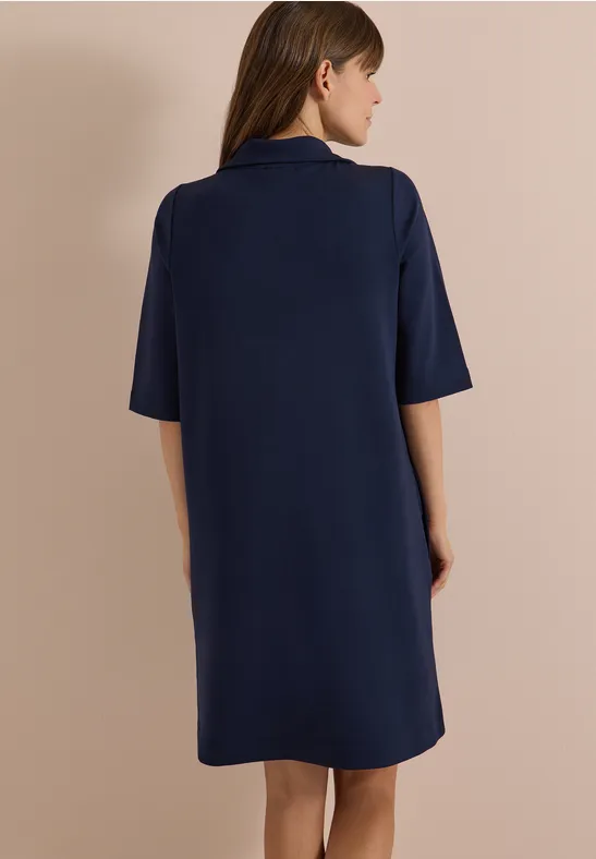 Une femme vue de dos porte une robe bleu marine ample arrivant au genou et aux manches mi-longues.