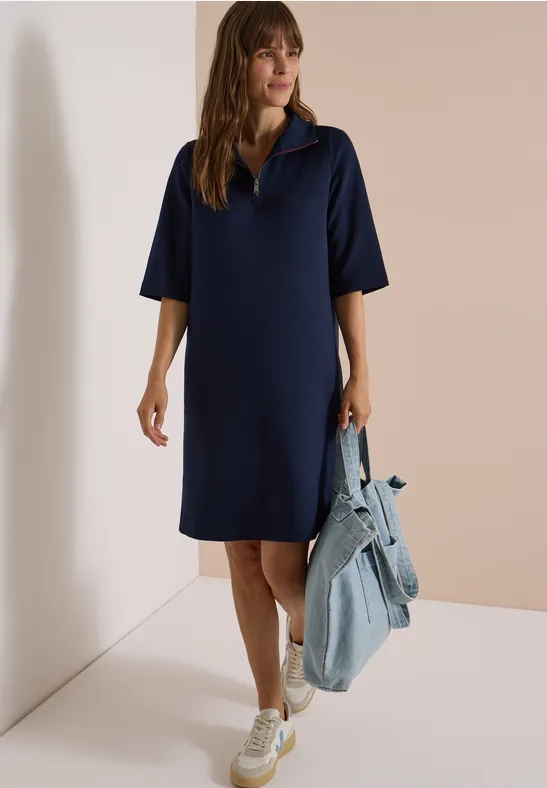 Femme en robe polo bleu marine ample jusqu'aux genoux, baskets blanches et veste en jean à la main.