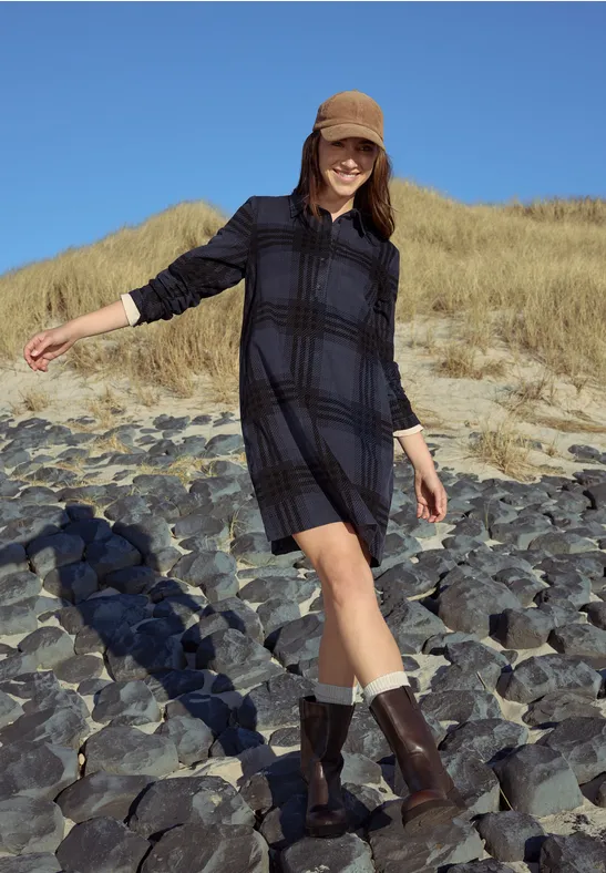 Femme en robe à carreaux, bottes et casquette marchant sur plage rocheuse près des dunes sous le soleil.