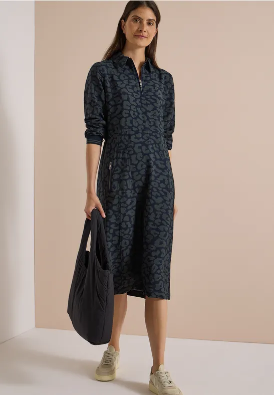 Femme portant une robe chemise bleu foncé à motif jusqu'aux genoux, manches retroussées, baskets beiges et sac noir.