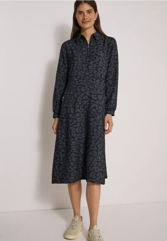 Femme en robe midi bleue à motif, col et manches longues, associée à des baskets blanches.