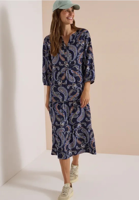 Femme en robe midi à motifs paisley bleu-blanc-rouge, manches 3/4, casquette, fond clair.