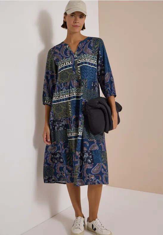 Femme en robe midi bleue à motifs, casquette blanche et baskets, tenant un sac noir devant un mur clair.