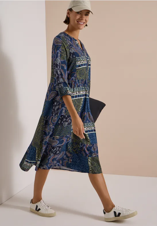 Femme souriante en robe bleue à motifs et baskets blanches, posant devant un fond clair.